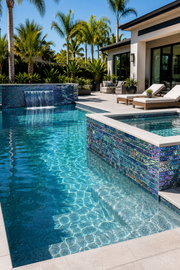 Pool with a raised wall and spillway tiled in Peacock Teal iridescent glass mosaic, beside a modern house and palms.