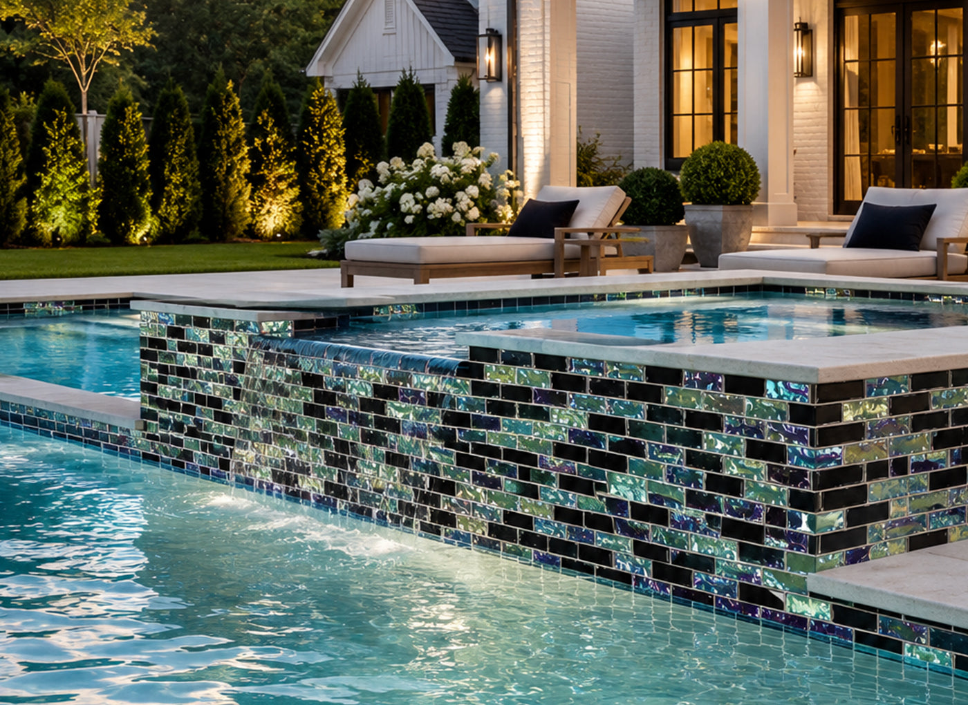 Pool at night with a Black Opal iridescent glass mosaic waterline band beside a lit house and hedge.