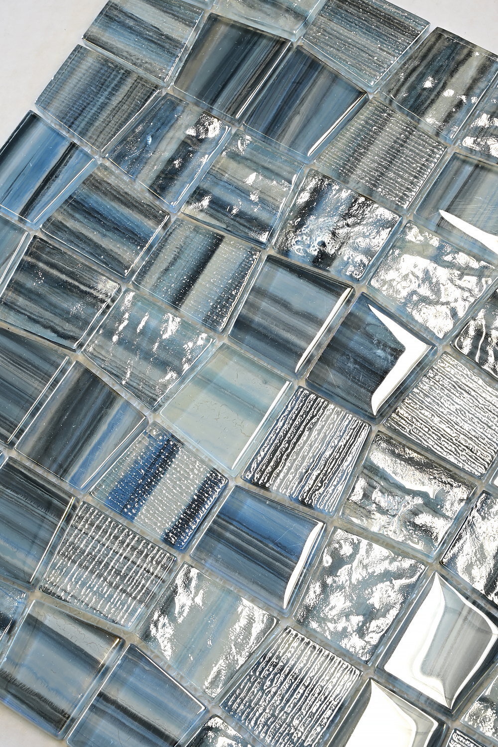 A close-up of glossy rectangular glass mosaic tiles in shades of blue, gray, and clear, arranged in a textured pattern.