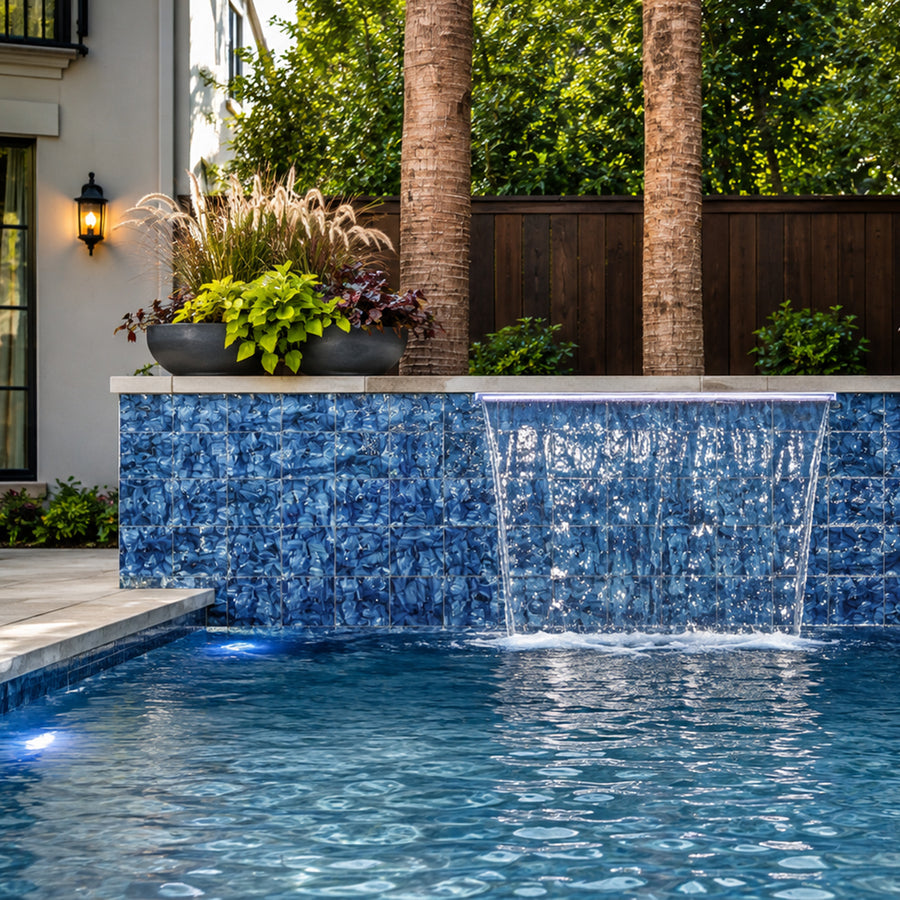 Waterfall spilling from a wall clad in Ocean Blue Artisan 6x6 porcelain tile beside lit garden planting.
