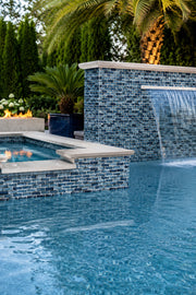 Pool with a waterfall wall clad in Storm Blue Marble recycled glass mosaic tile, framed by palms and planters.