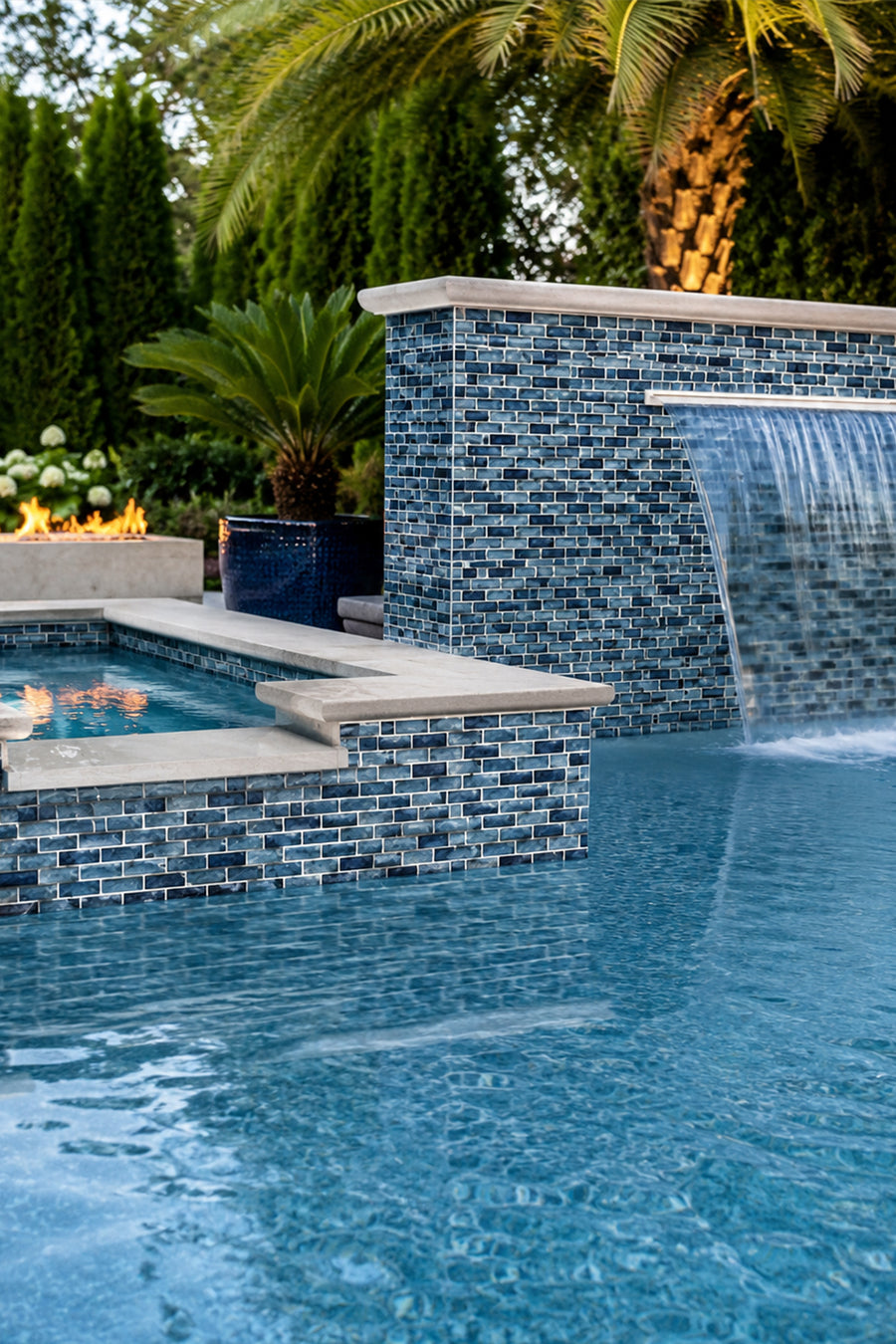 Pool with a waterfall wall clad in Storm Blue Marble recycled glass mosaic tile, framed by palms and planters.